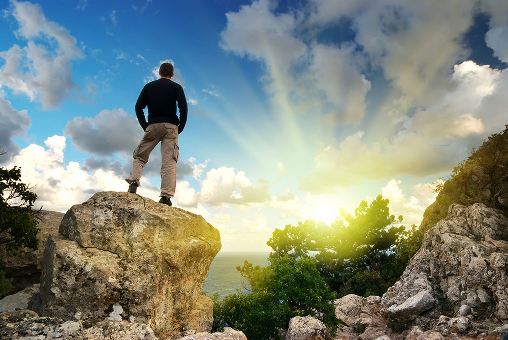 Man standing tall on a large boulder overlooking a sunny distance