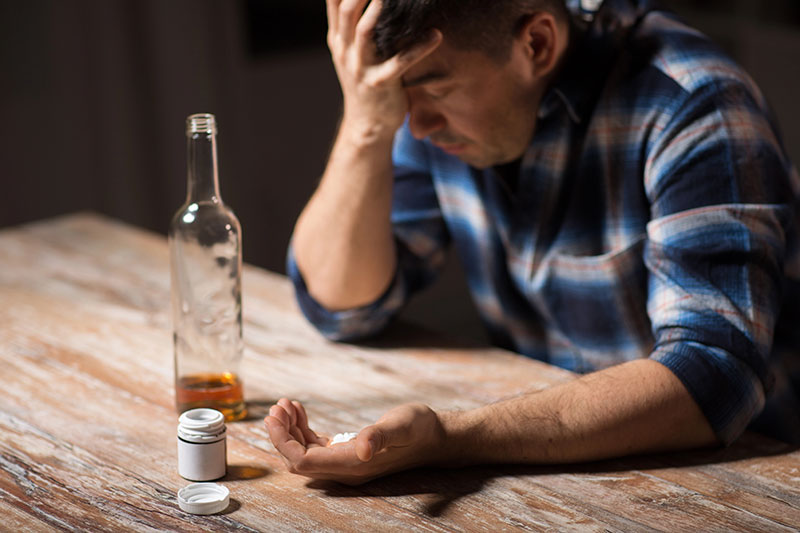 Man with his head in his palm, pills in his other hand, an almost empty bottle of alcohol and a pill bottle on the table.